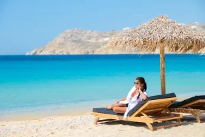Woman relaxing on a beach chair under a straw umbrella in Mykonos 2025 with turquoise sea in the background