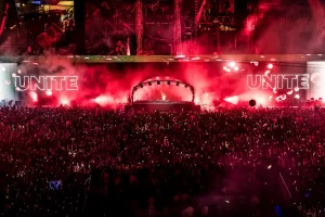 DJ performing on the Unite mainstage at Tomorrowland Belgium 2025 under red lights, with a crowd of festivalgoers holding up phones